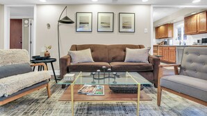 Un salon moderne avec un canapé marron, une table basse en verre et un fauteuil gris. Des tableaux encadrés ornent le mur, ainsi qu’une lampe sur pied et des meubles en bois dans l’espace cuisine.