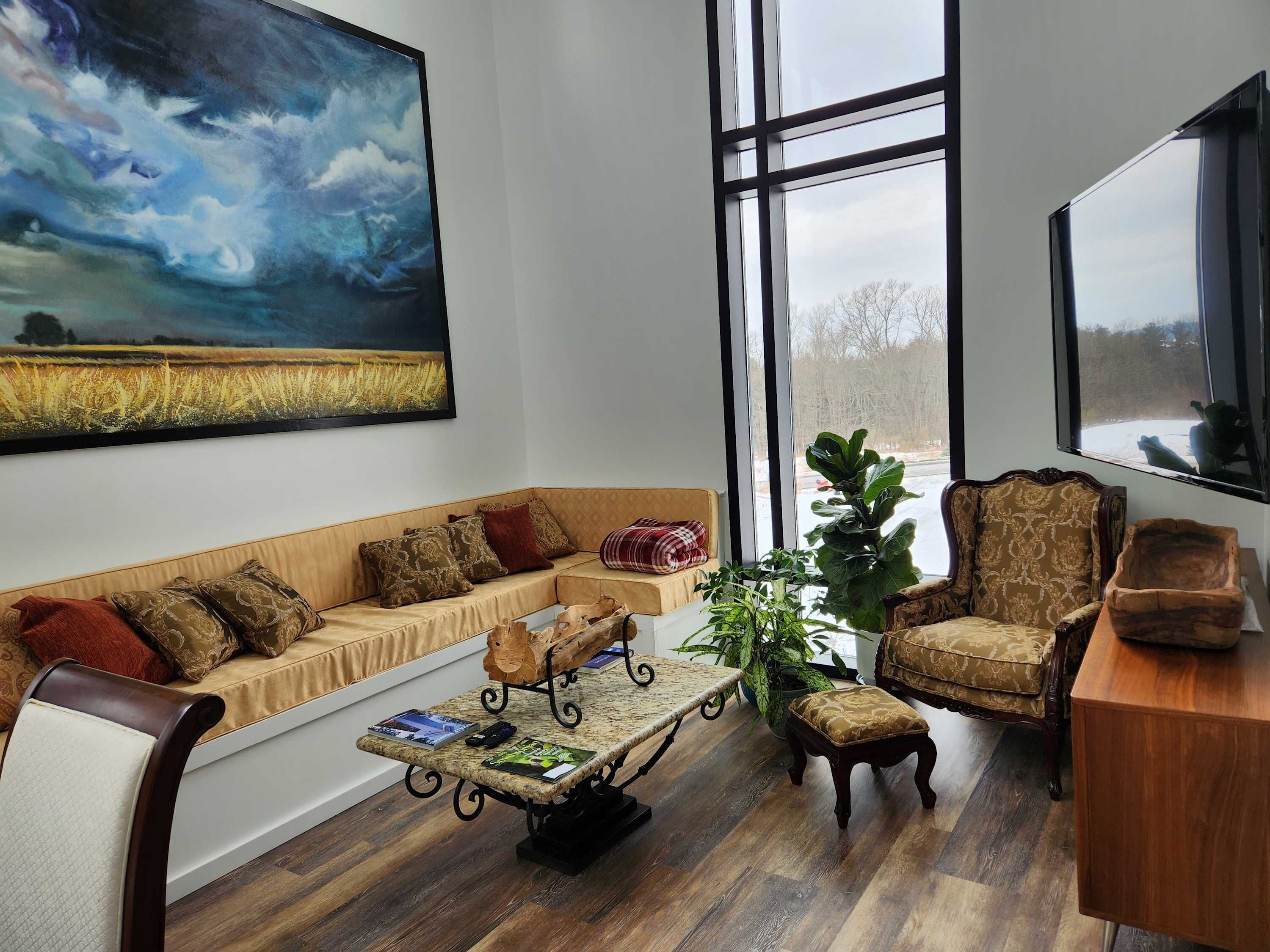 Un salon chaleureux avec un grand tableau, un canapé, un fauteuil, une table basse et une grande fenêtre.