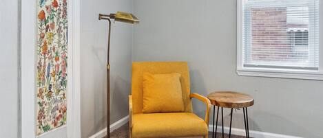 Ein Zimmer mit einem senfgelben Sessel, einem kleinen Holzschemel, einer hohen Stehlampe und einem Wandteppich mit Blumenmuster.