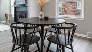 A round dining table with dark wooden chairs, a vase with dried plants, and a flat-screen TV above a fireplace. - Plainview Haven (Detroit)