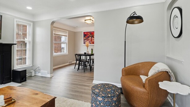 A living room with a brown leather sofa, a round ottoman, a wooden coffee table, a black floor lamp, and a dining area in the background. - Plainview Haven (Detroit)