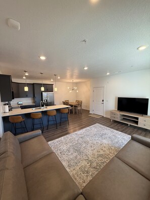 A modern living room with a sofa, a TV, and a kitchen area with bar stools. - Brand New Condo in Spanish Fork (Spanish Fork)