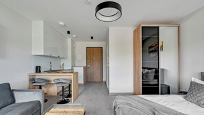 A modern hotel room with a grey sofa, a wooden coffee table, a bed with a grey blanket, a desk with two chairs, a wardrobe, and a door leading to a bathroom.