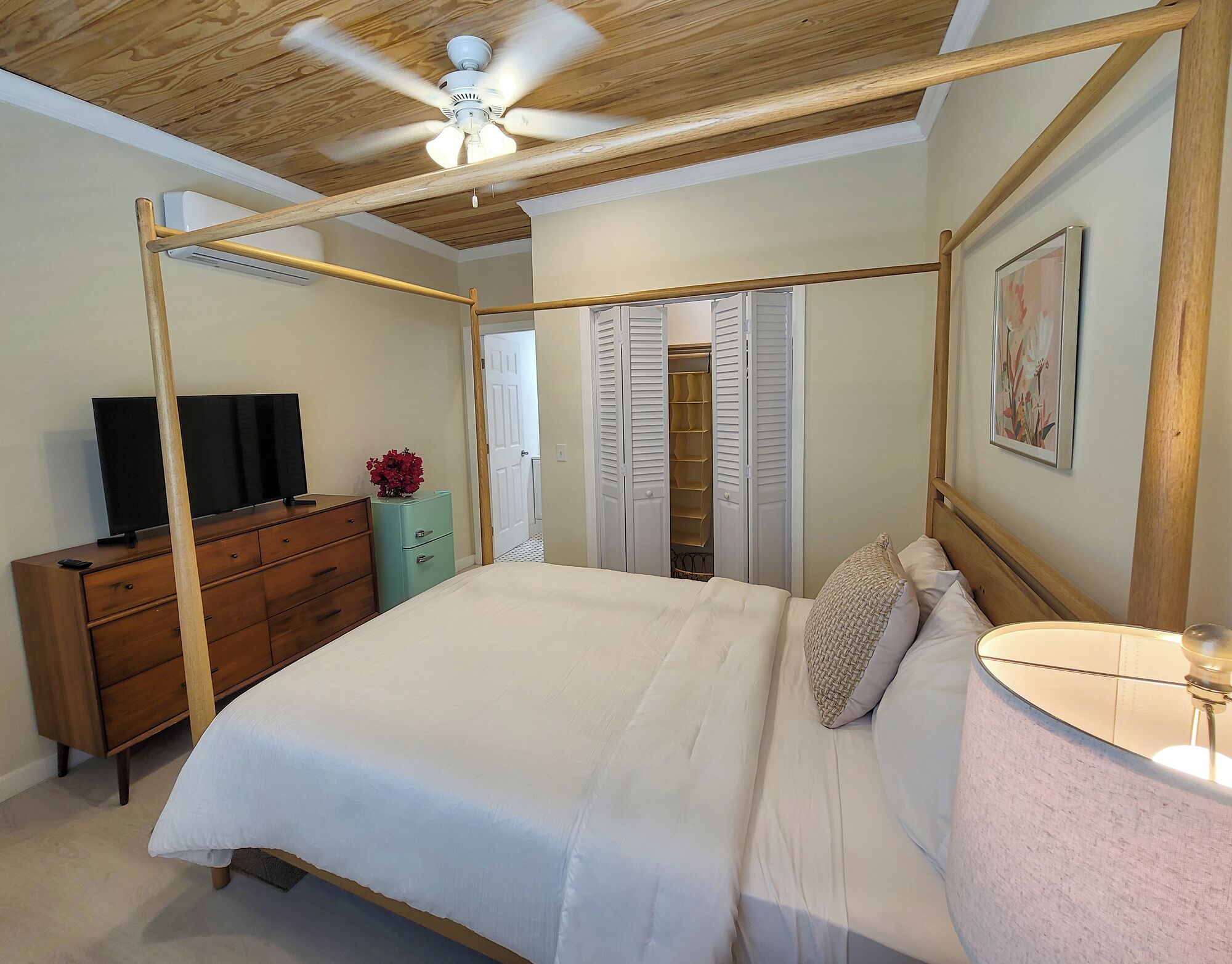 Un dormitorio con cama con dosel, ventilador de techo, televisor sobre un aparador de madera y armario con puertas correderas.