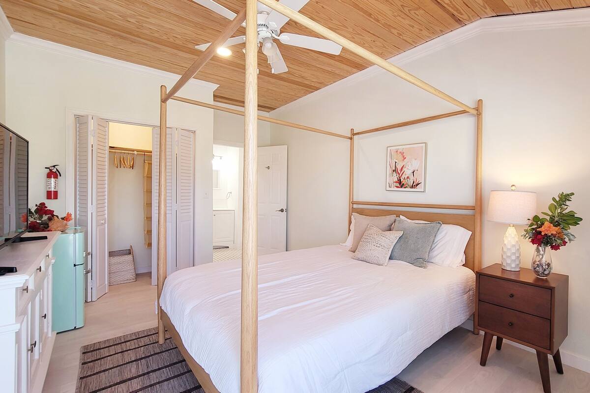 Ein Schlafzimmer mit einem Himmelbett, einem Nachttisch mit Lampe und Blumen, einem Deckenventilator und einem an der Wand montierten Feuerlöscher.