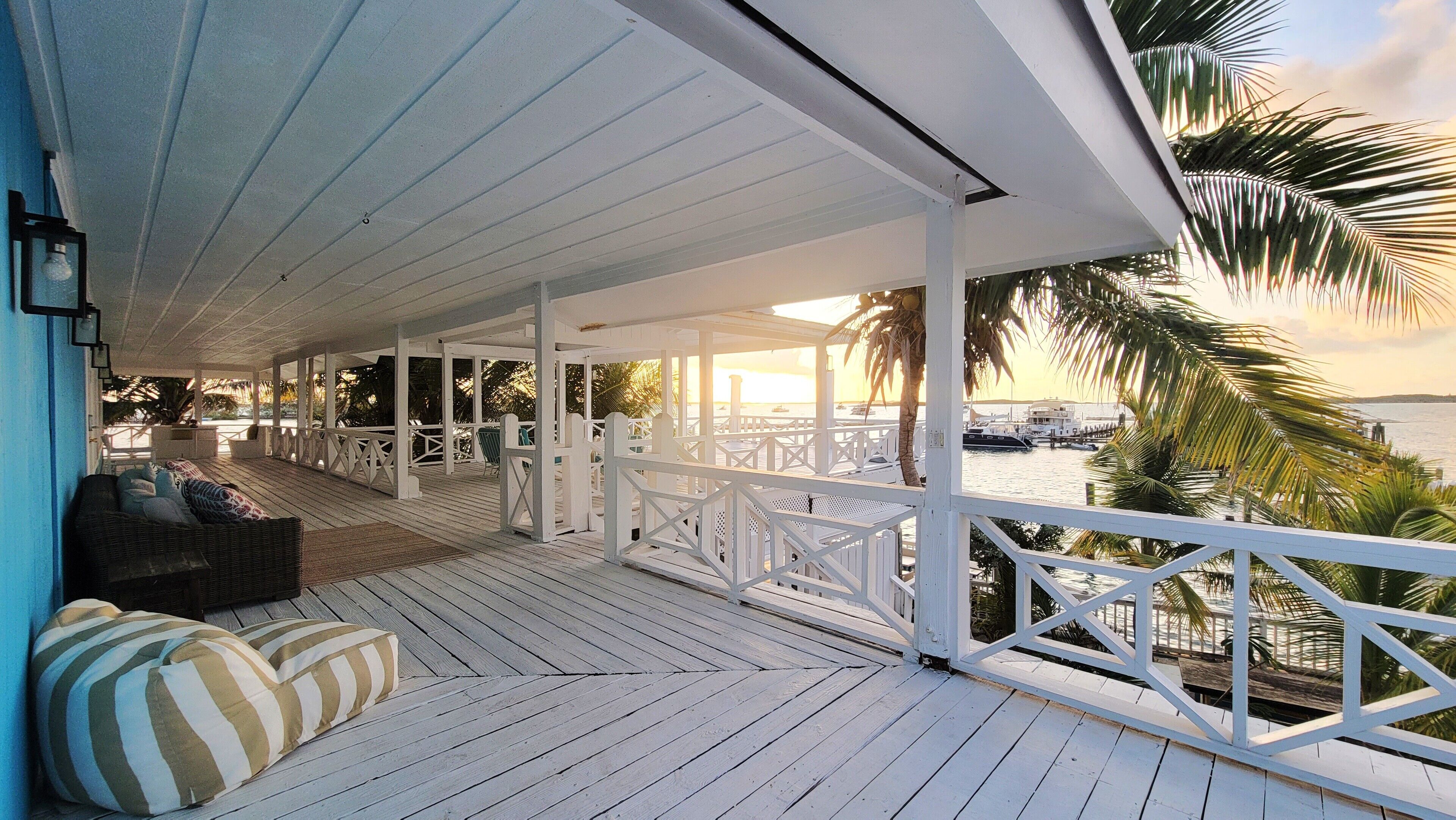 A covered wooden deck with white railings, a wicker sofa with striped cushions, and a view of the sea and boats at sunset.