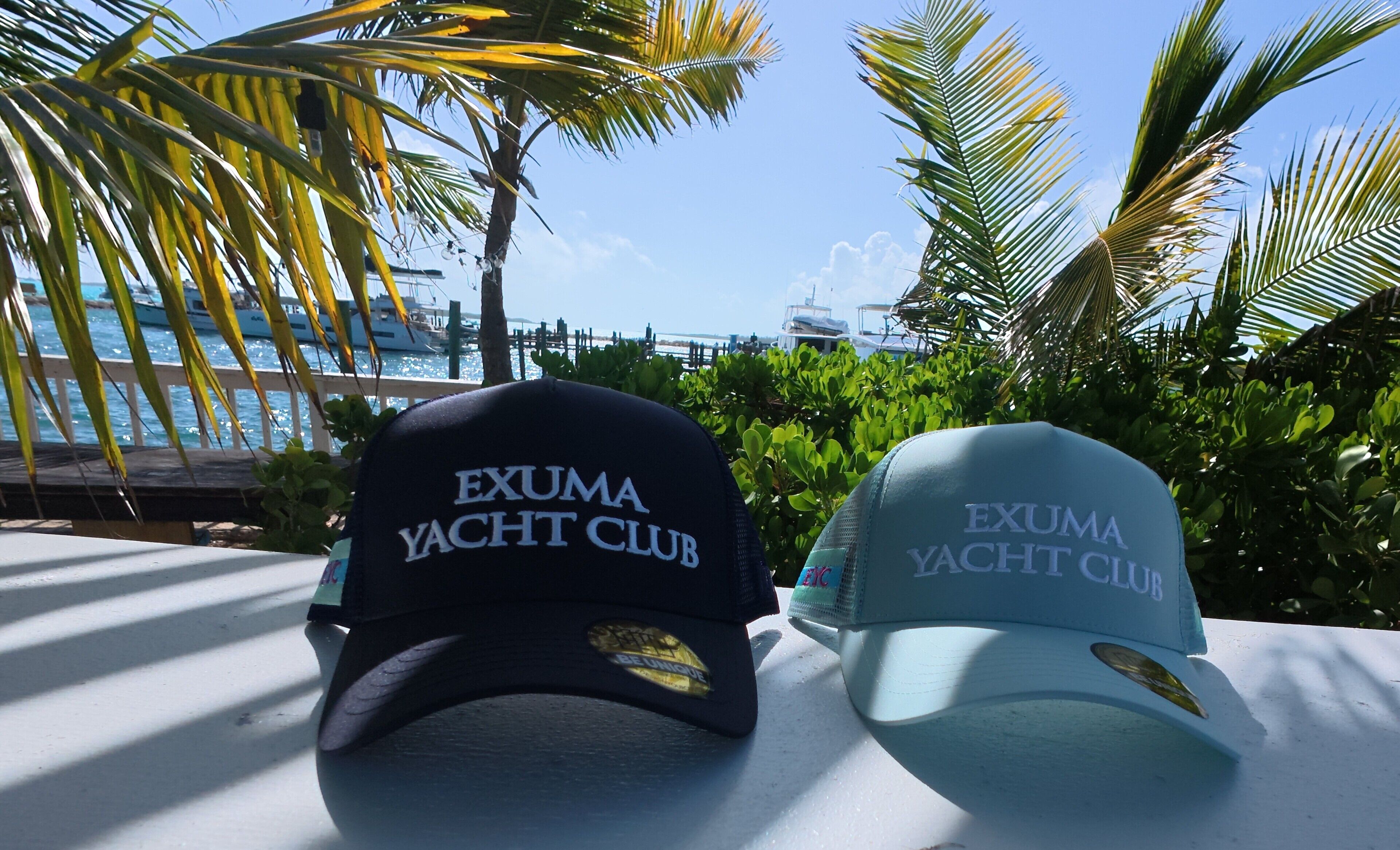 Deux casquettes ornées de l’inscription « Exuma Yacht Club », posées sur une table offrant une vue sur la mer et les palmiers.