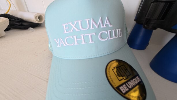 A light blue cap with embroidered text "EXUMA YACHT CLUB" and a gold emblem with "BE UNIQUE" on a white background. - Exuma Yacht Club Resort & Marina Limited (George Town)