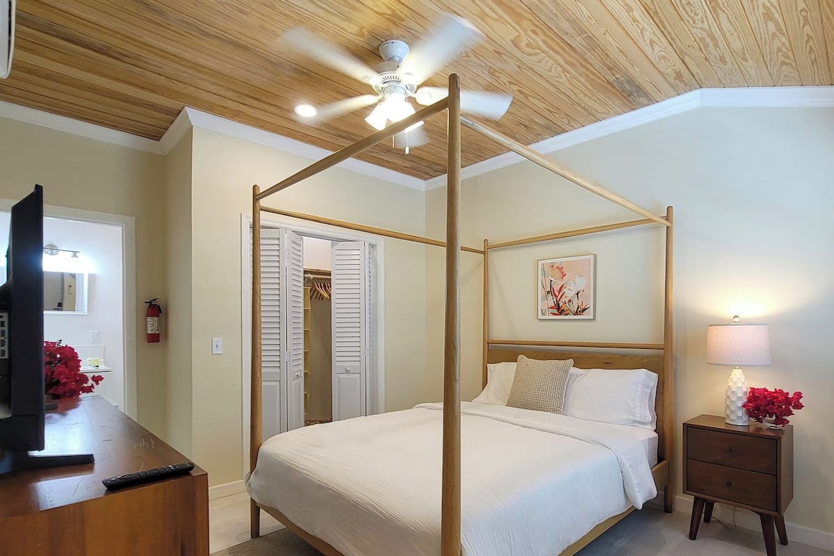 Ein Schlafzimmer mit einem Himmelbett, einem hölzernen Deckenventilator, einem Nachttisch mit Lampe und einem Bild an der Wand.