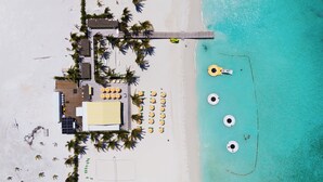 Vista aérea de un resort frente a la playa con zona de piscina, tumbonas y un muelle que se adentra en el mar turquesa.