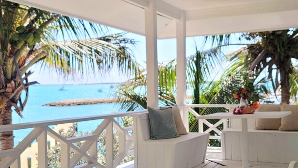 Un área de estar exterior techada con mobiliario blanco, vista al océano y un jarrón con flores rojas.