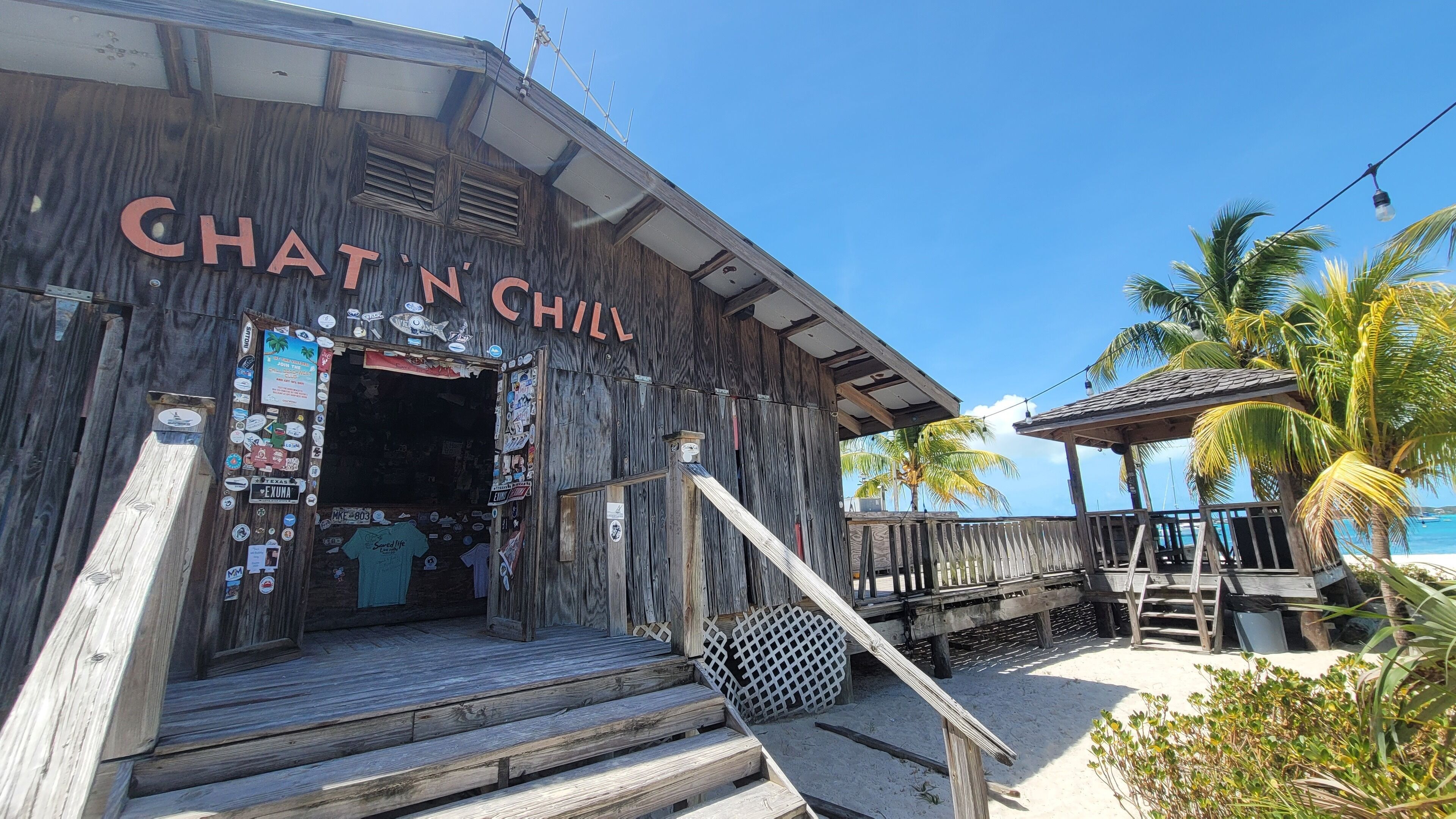 A wooden building with a sign reading "CHAT N' CHILL" and a beachside view with palm trees and a wooden deck.