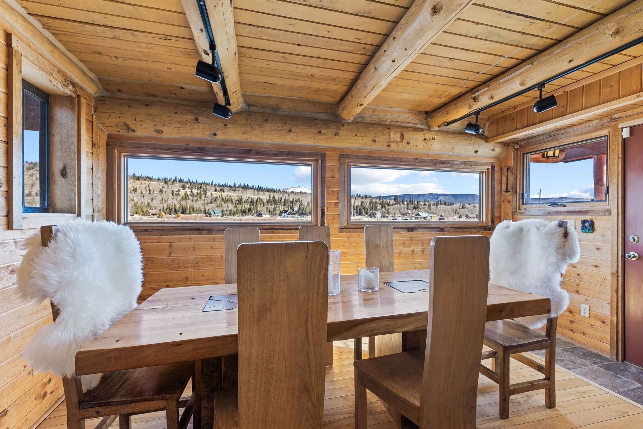 Interior de una cabaña de madera con una mesa y sillas de comedor, ventanas amplias con vista a un paisaje boscoso y una puerta de madera.