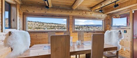 Interior de una cabaña de madera con una mesa y sillas de comedor, ventanas amplias con vista a un paisaje boscoso y una puerta de madera.