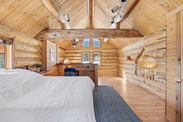 A log cabin bedroom with a large bed, a desk, and a chair. There are two windows and a ceiling fan.
