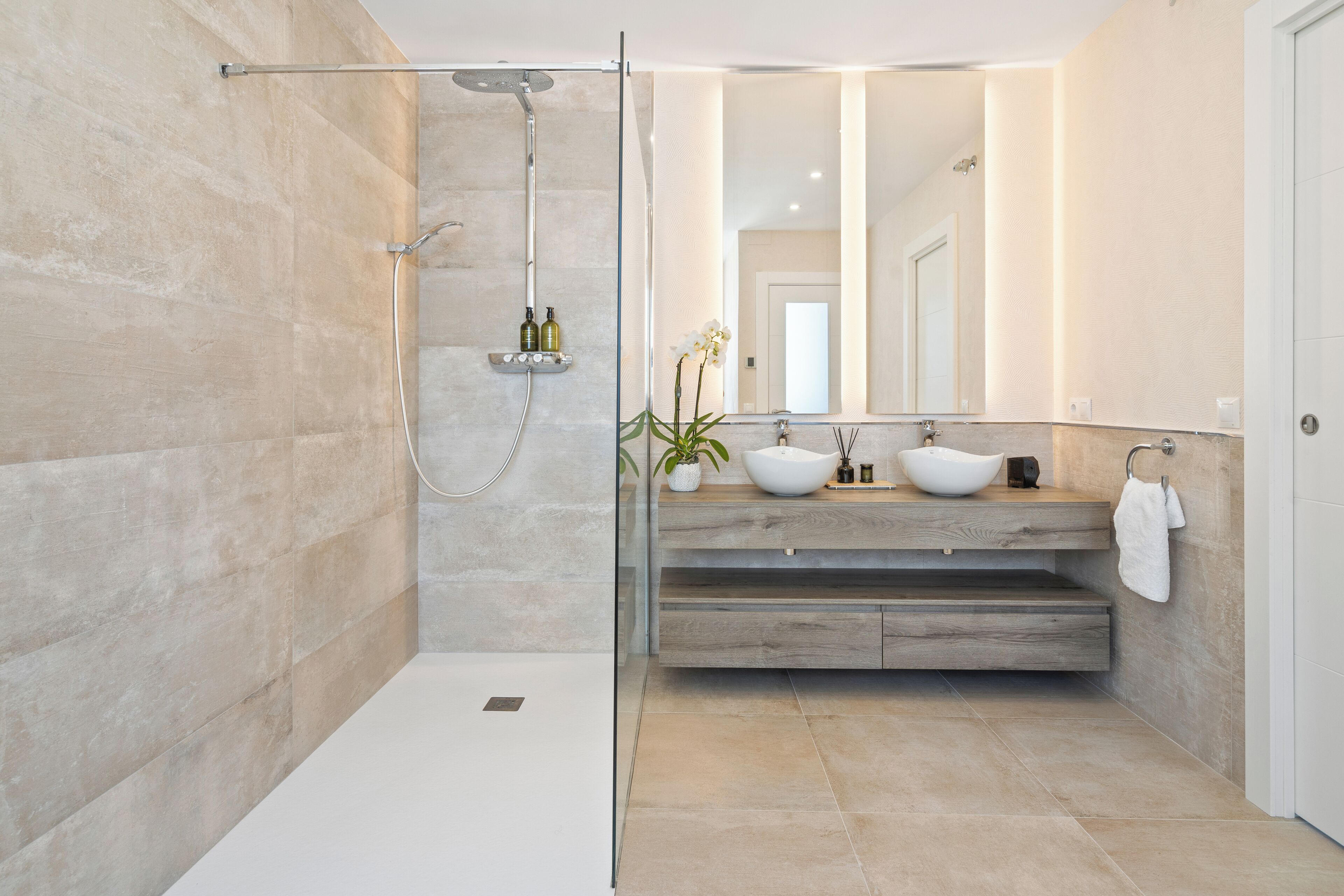 Un baño moderno con ducha envuelta en vidrio, tocador de madera con lavabo blanco y un espejo grande.