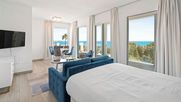 Ett modernt hotellrum med en stor säng, en TV, en matplats med bord och stolar samt en balkong med utsikt över havet.