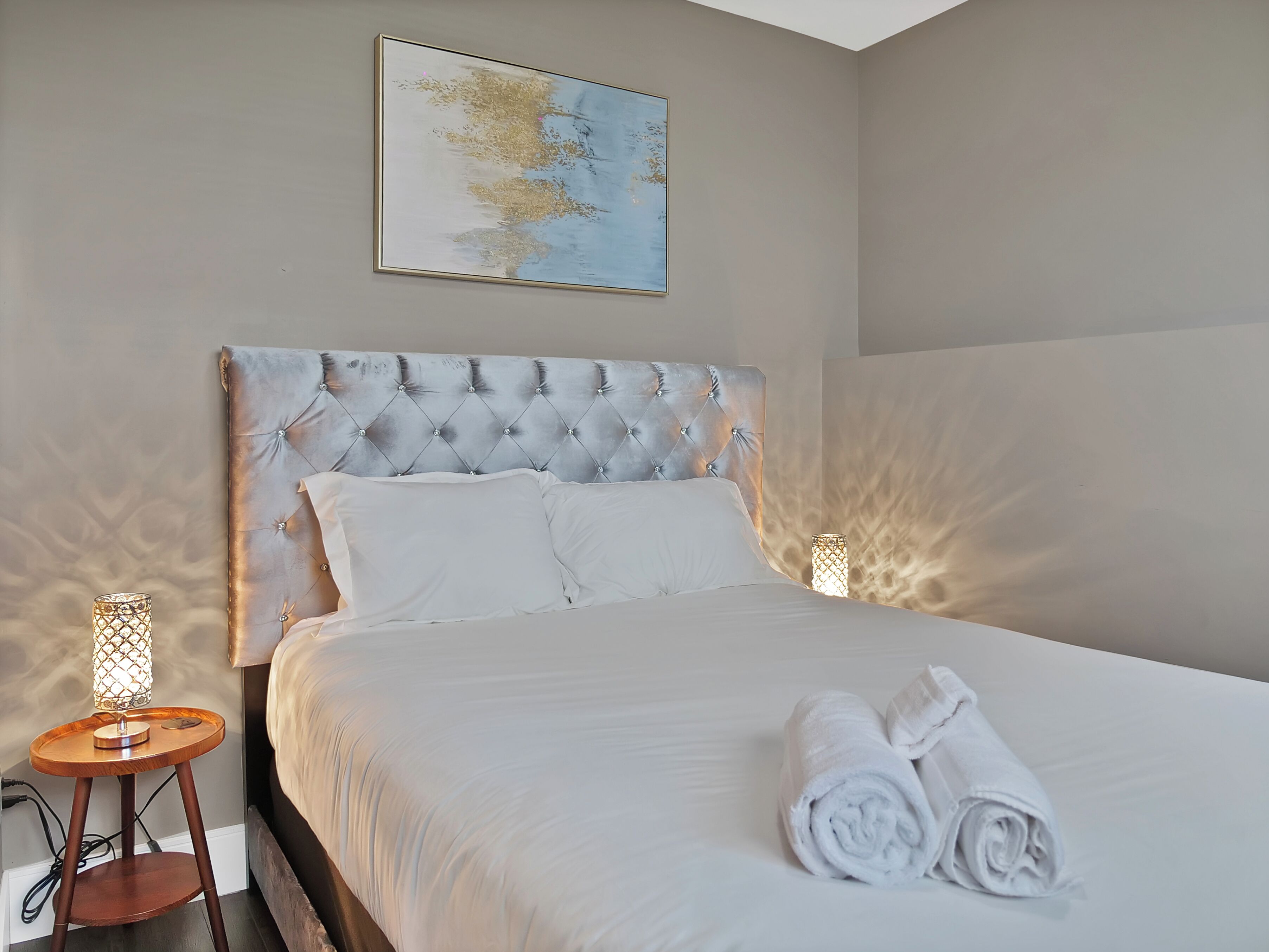 Een netjes opgemaakt bed met witte lakens en opgerolde handdoeken, een gestuikt hoofdbord en twee bedlampen op houten nachtkastjes.