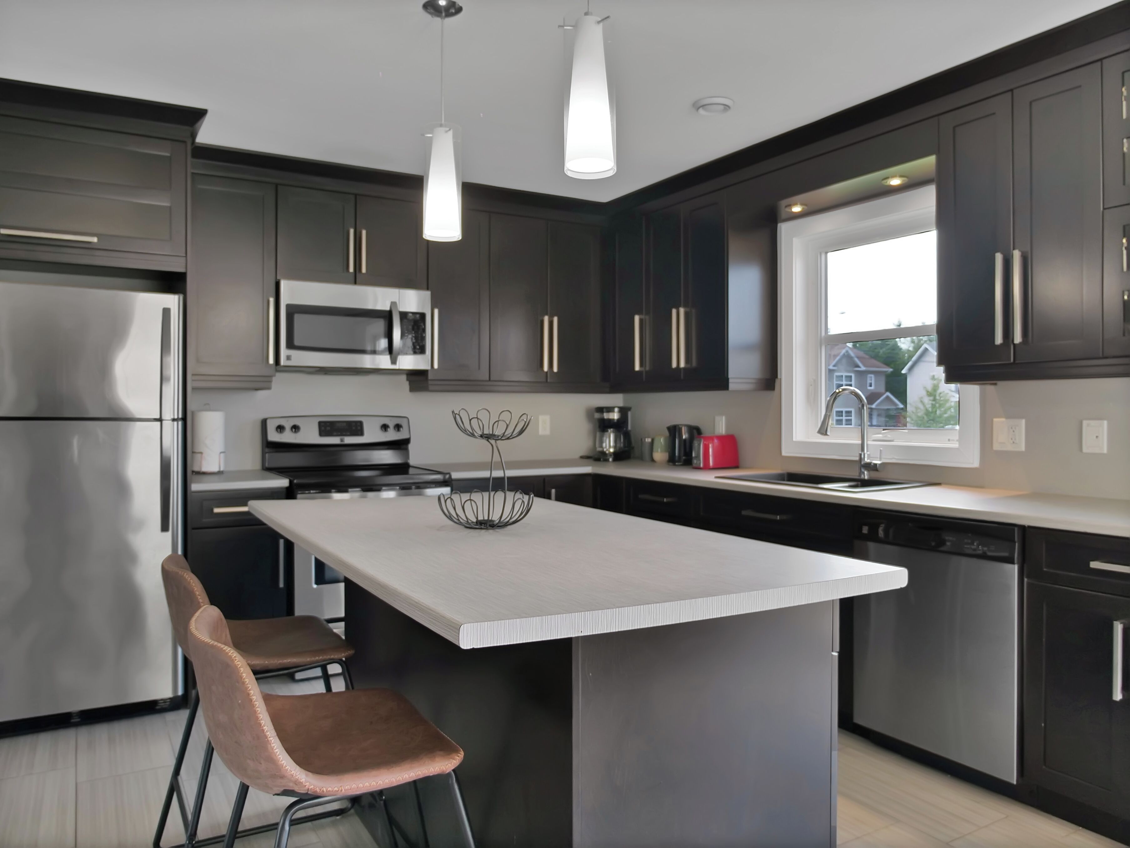Une cuisine moderne avec des meubles de couleur sombre, des appareils électroménagers en acier inoxydable, un îlot central et des luminaires suspendus.
