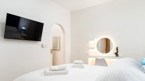 Et hotelværelse med en seng, et fladskærms-tv monteret på væggen, et rundt spejl med lys og en dør med buet indgang.