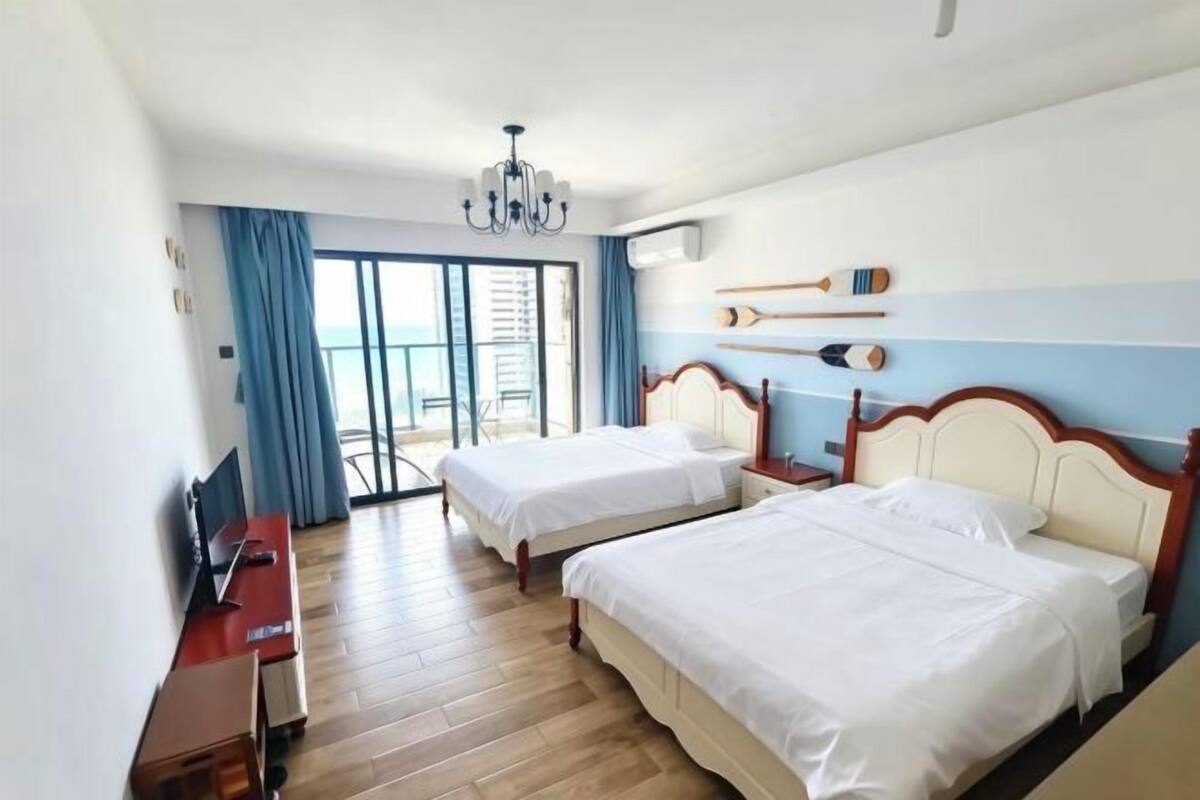 A hotel room with two beds, a desk, a TV, a chandelier, and a view of the sea through a sliding door.