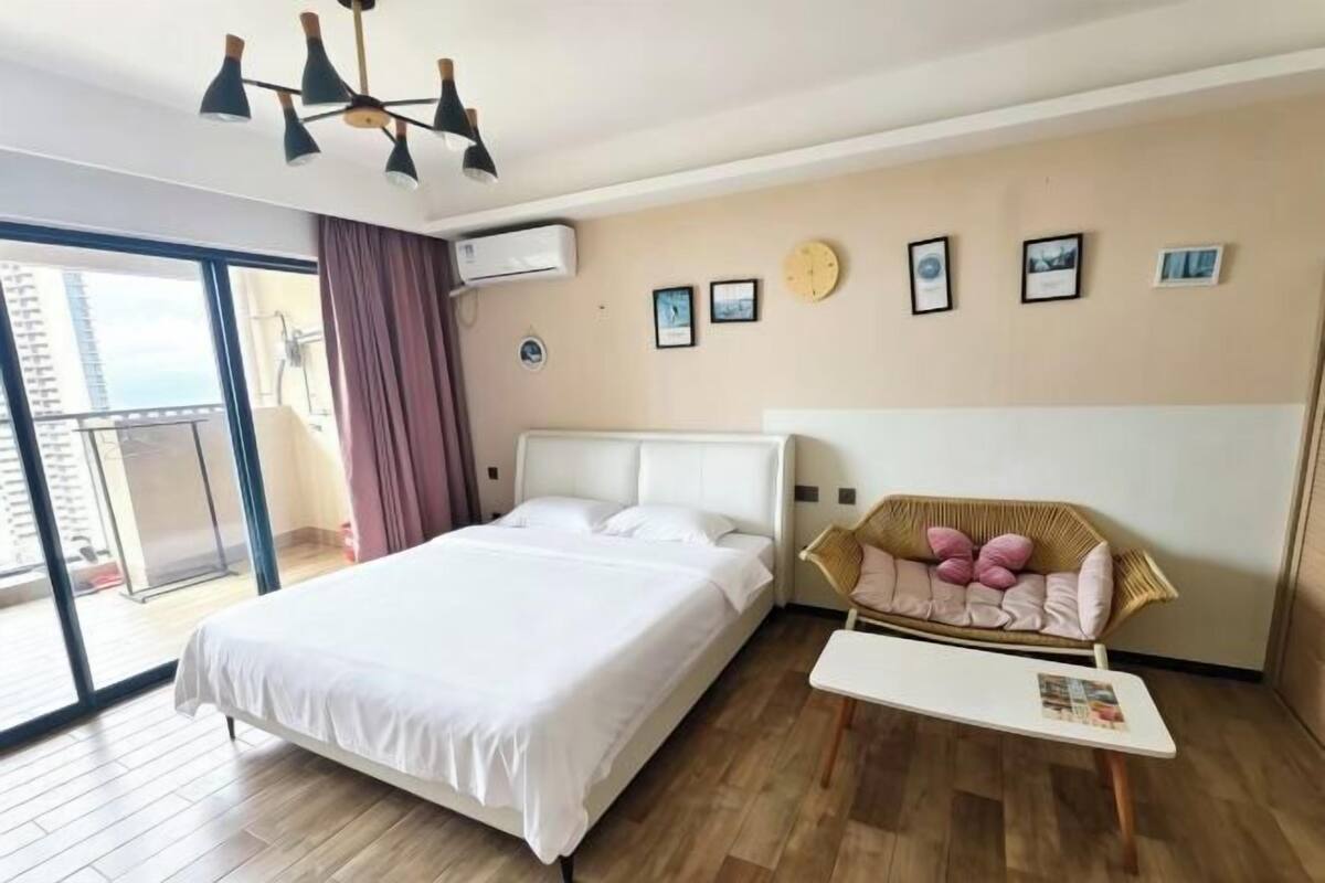 A hotel room with a bed, a small table, a wicker chair, a balcony, and a wall with framed pictures.