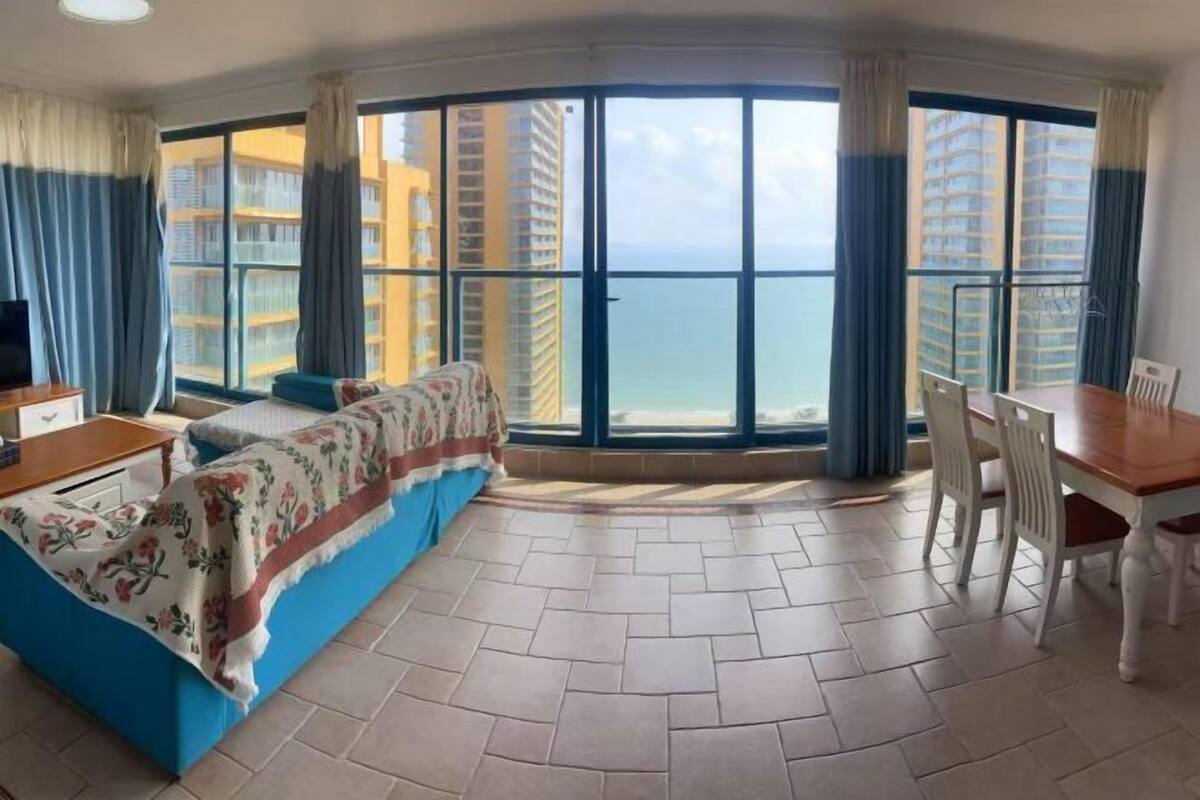A corner room with a bed, a dining table, and a view of tall buildings and the sea.