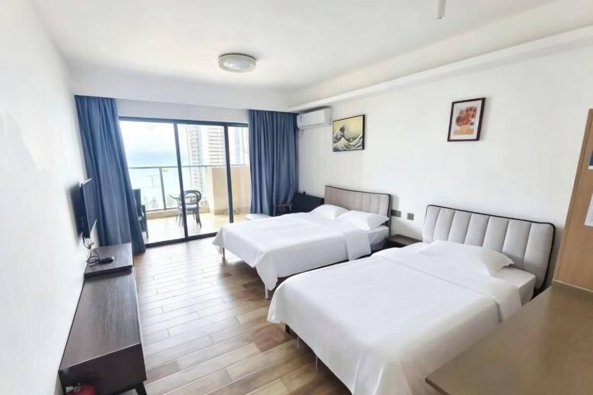 A hotel room with two beds, a desk, a chair, and a balcony with a view.