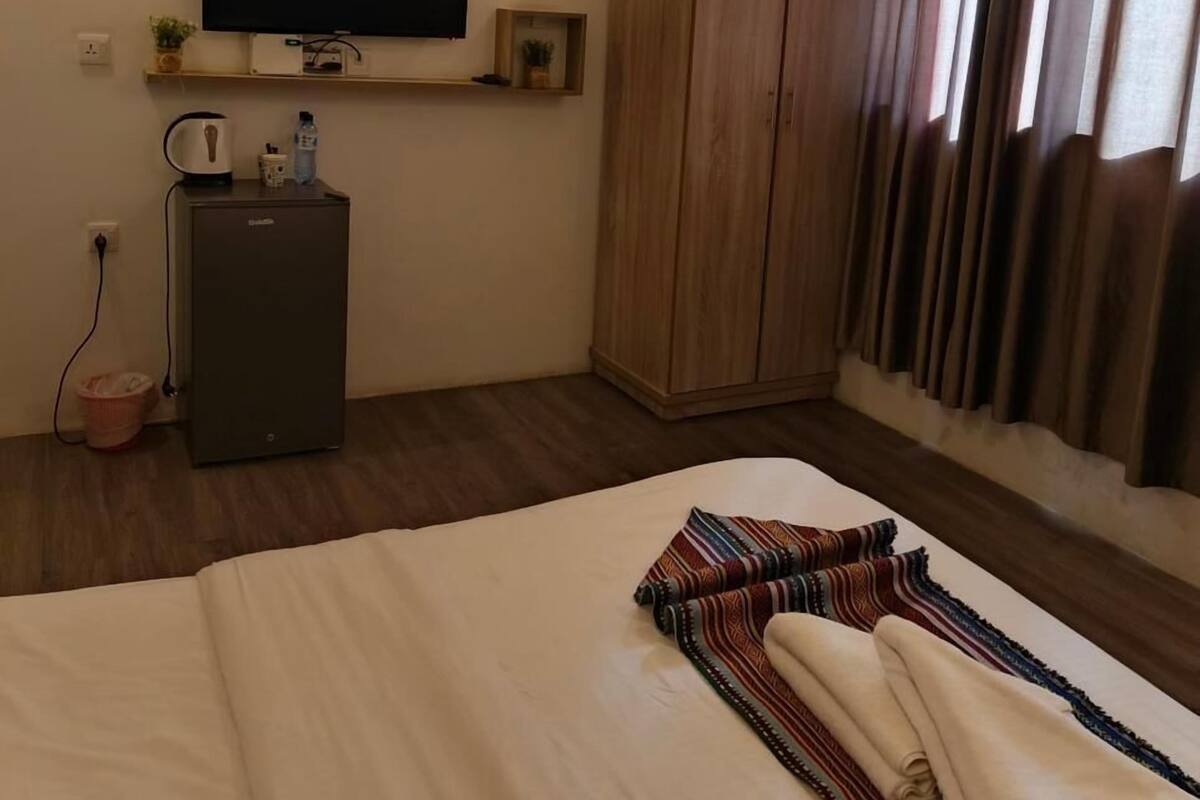 A hotel room with a bed, a TV mounted on the wall, a wardrobe, a small fridge, and a window with curtains.