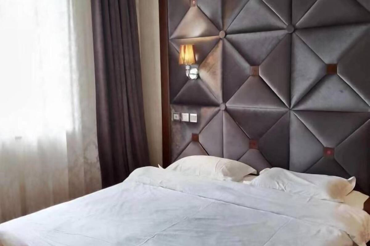 A hotel room with a bed, a wall-mounted light, and a window with curtains.