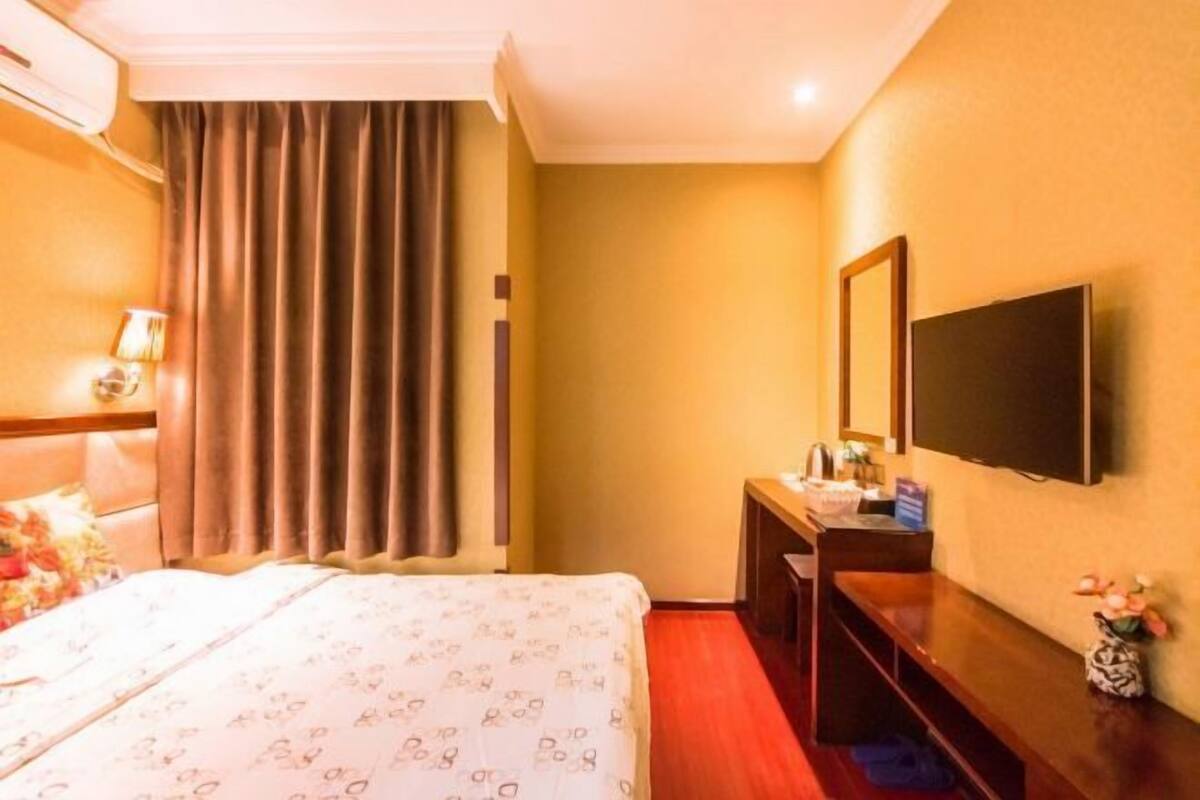 A hotel room with a bed, a TV mounted on the wall, a desk with a chair, and a window with curtains.