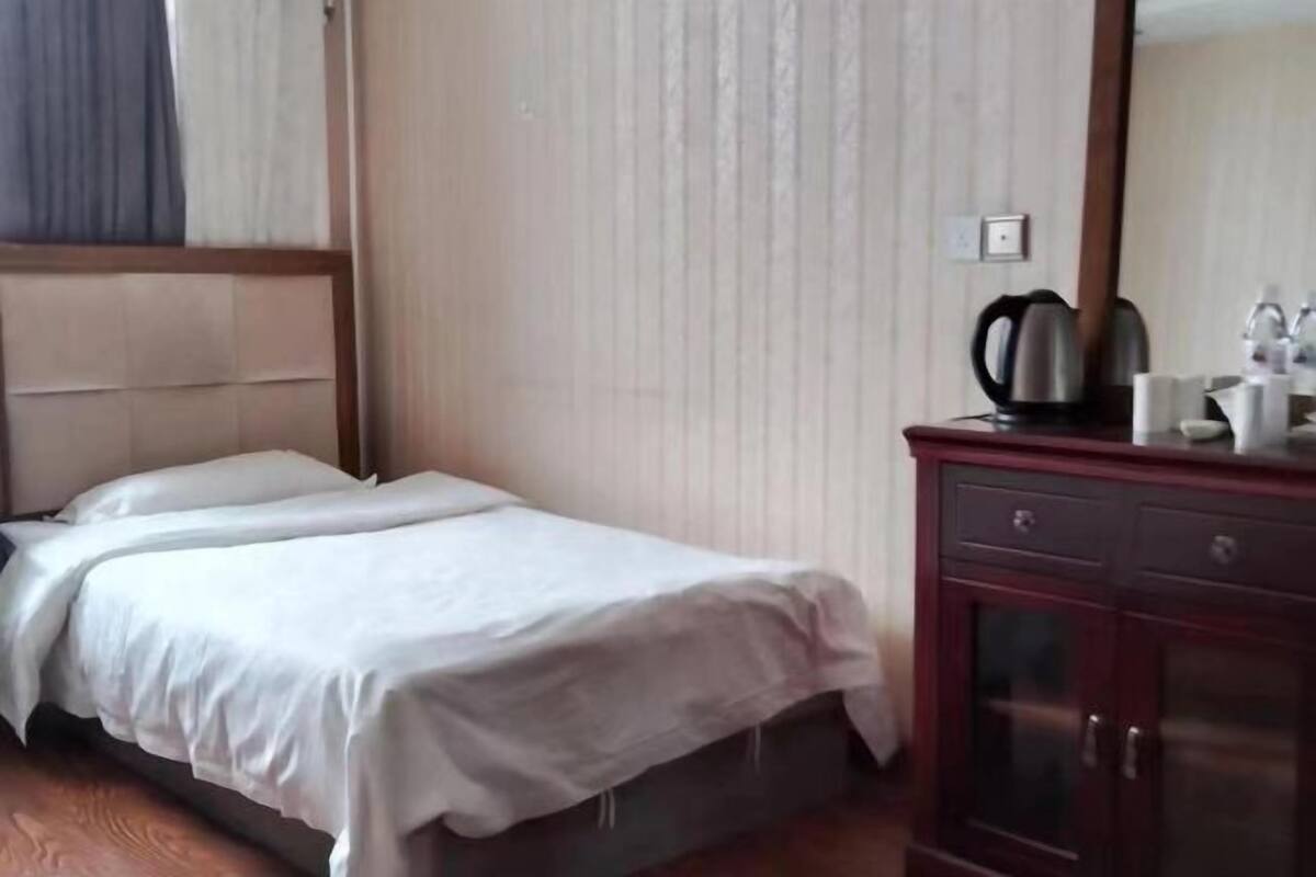 A hotel room with a bed, a wooden headboard, a dark wooden dresser with a mirror, and a kettle on the dresser.