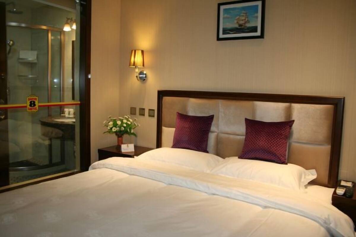A hotel room with a bed, a nightstand, a lamp, a picture on the wall, and a glass door leading to a bathroom.