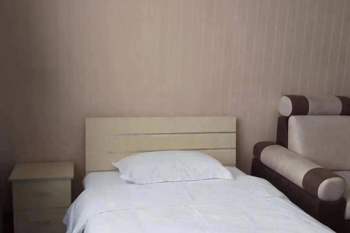 A neatly made bed with a white pillow and a wooden headboard, a nightstand to the left, and a brown armchair to the right.