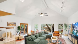 A spacious living room with a large sectional sofa, a coffee table, and a fireplace. There are multiple windows providing natural light and views of the outdoors. - Ultimate Retreat 5-bed, Pool, Hot Tub, Mini Golf, Game Room, Playground and More (San Antonio)