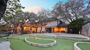 A courtyard with a putting green, stone borders, and a building with string lights. - Ultimate Retreat 5-bed, Pool, Hot Tub, Mini Golf, Game Room, Playground and More (San Antonio)