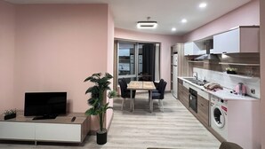 Una sala de estar moderna con televisor de pantalla plana, un sofá, una mesa de comedor con sillas, un área de cocina con armarios y electrodomésticos, y una planta en maceta.