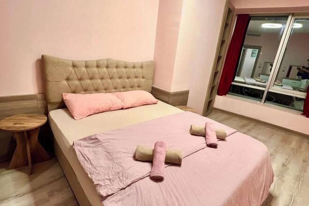 A bedroom with a tufted headboard, a bed with pink bedding, two rolled towels, a small wooden side table, and a window with a red curtain.