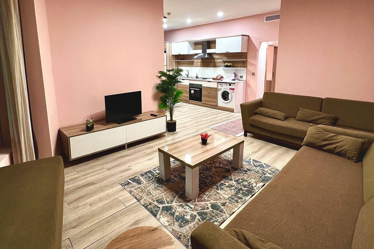 A living room with a sofa, a coffee table, a TV, and a kitchen in the background.