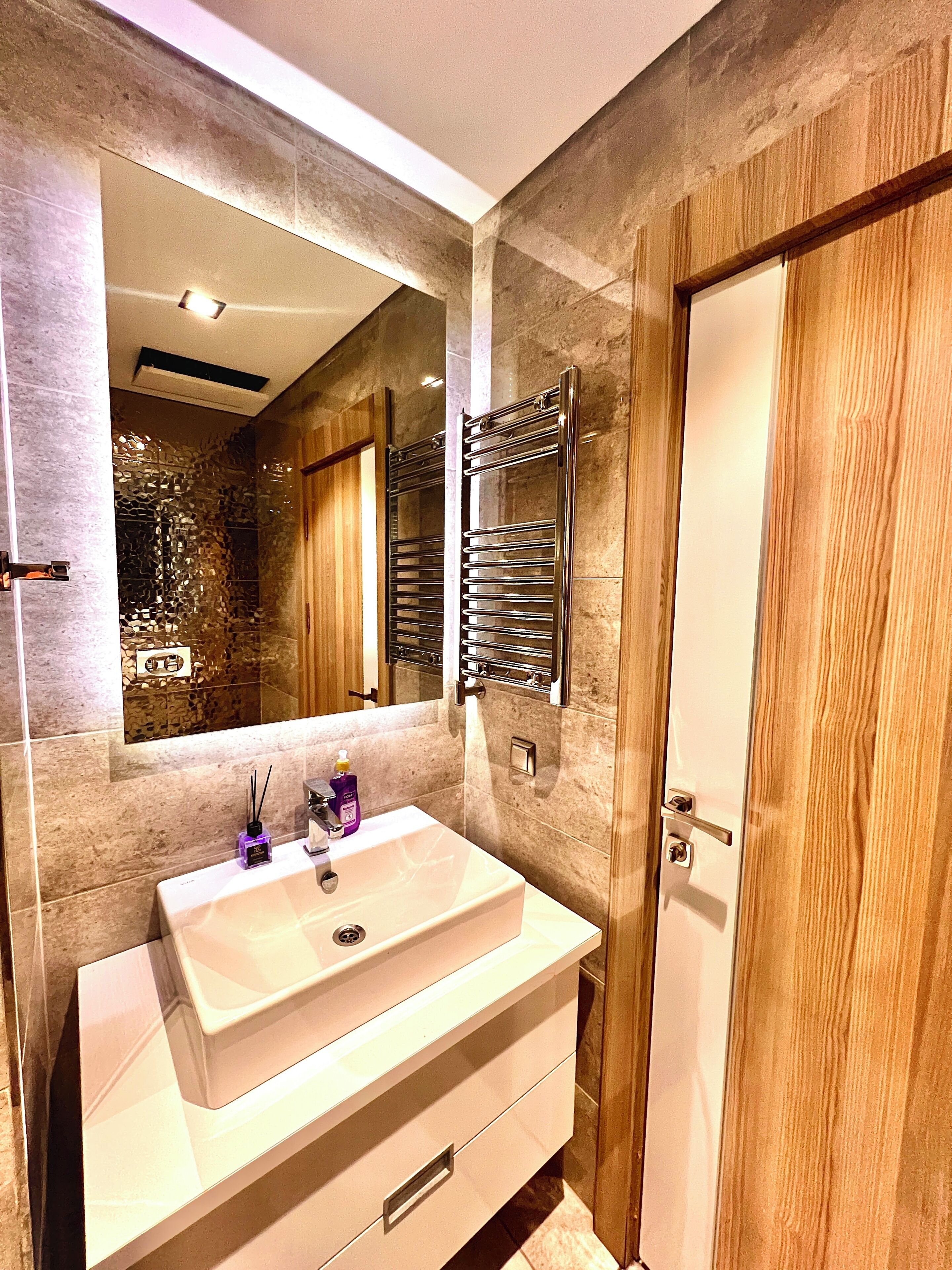 A modern bathroom with a large mirror, a white sink, a towel rack, and a wooden door.