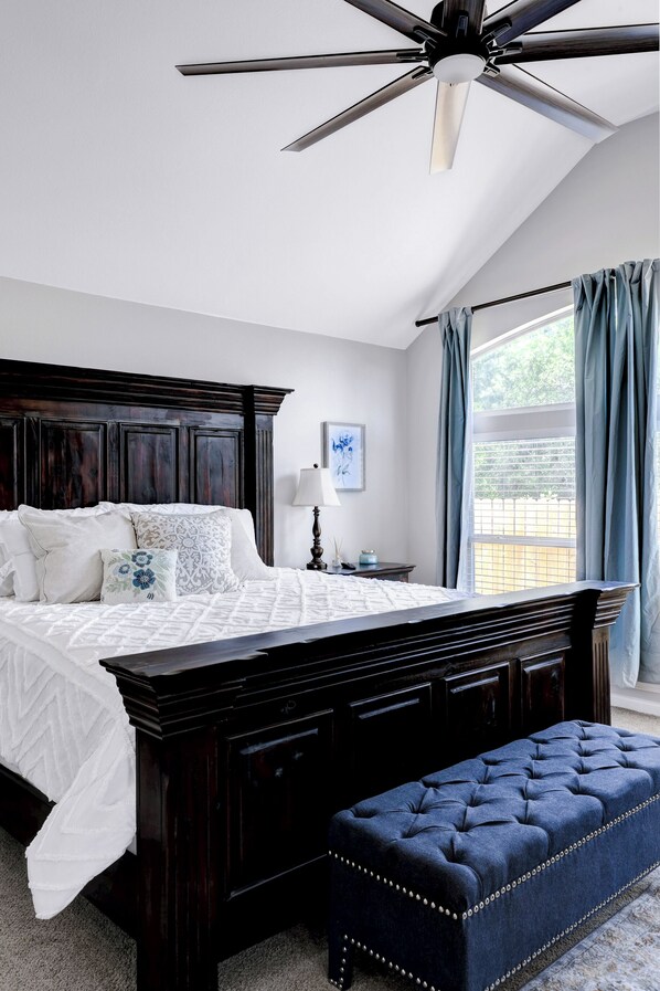 Un dormitorio con una cama de madera oscura, un ventilador de techo, una ventana con cortinas y una mesita de noche con lámpara.