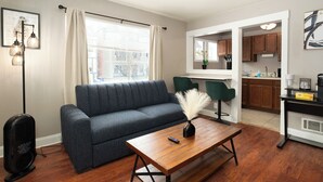 Una sala de estar con un sofá, una mesa de centro y un televisor. Se ve un área de cocina a través de una puerta abierta.