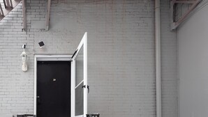 Pared de ladrillo blanco con una puerta negra, una cámara de seguridad y una pequeña mesa con un teléfono.