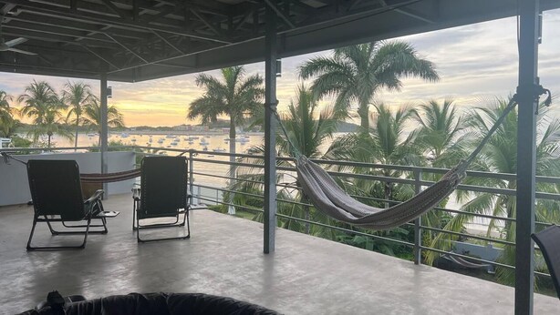 A balcony with two chairs and a hammock overlooking a waterfront with palm trees and boats. - Beachfront 2nd level townhome. (Santa Cecilia)