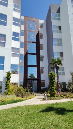 A modern multi-story building with a central glass facade, surrounded by well-maintained landscaping and palm trees. - 3BR Oceanview Penthouse, AC, Sleep 8, Gated, Pool, Playground, BBQ, Security24-7 (Rosarito)