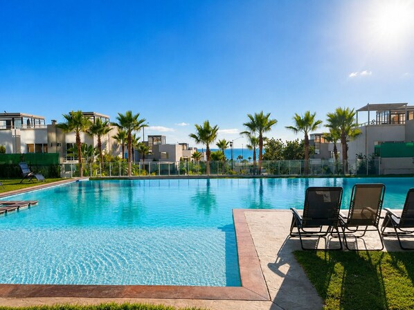 A swimming pool with lounge chairs, palm trees, and a view of the ocean. - 3BR Oceanview Penthouse, AC, Sleep 8, Gated, Pool, Playground, BBQ, Security24-7 (Rosarito)