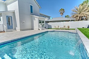 A swimming pool with a clear blue water, surrounded by a white house with a blue door and windows, and a white fence.
