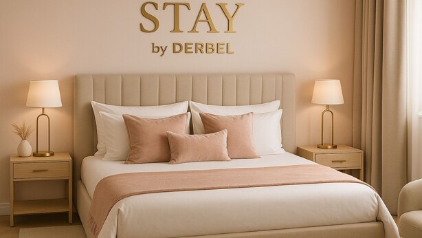 A hotel room with a large bed, two bedside tables with lamps, a wall-mounted sign reading "STAY by DERBEL", and a window with curtains.