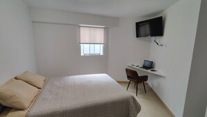 Een kleine, netjes ingerichte slaapkamer met een bed, een bureau met een laptop, een stoel en een aan de muur bevestigde televisie.