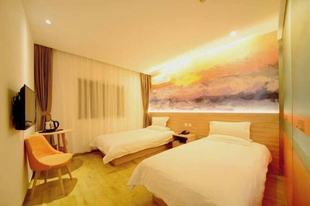 A hotel room with two beds, a desk, a chair, and a large window with a sunset view.